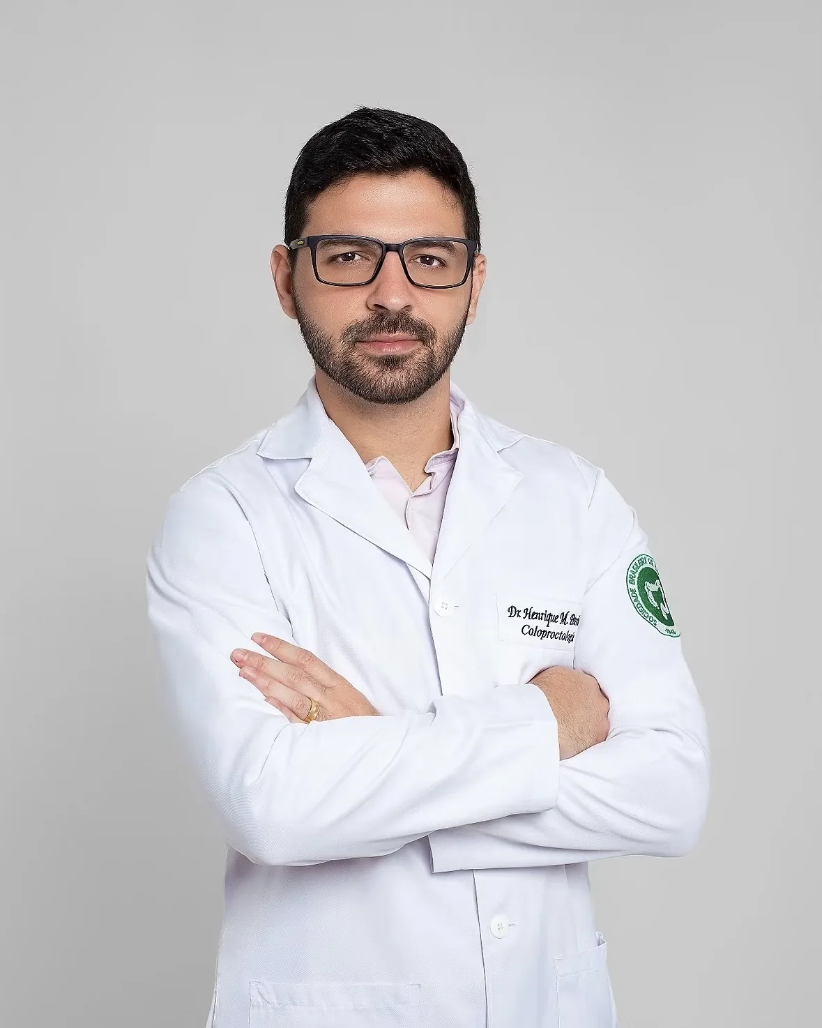 Dr. Henrique Moura, de jaleco com a identificação de coloproctologista, braços cruzados, sobre fundo cinza claro