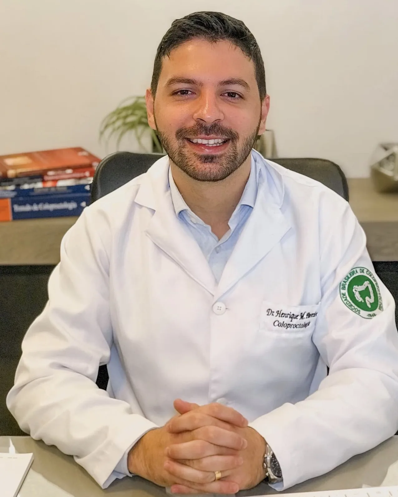 Dr. Henrique Moura sentado à mesa do consultório, de jaleco, sorrindo, com livros de coloproctologia ao fundo